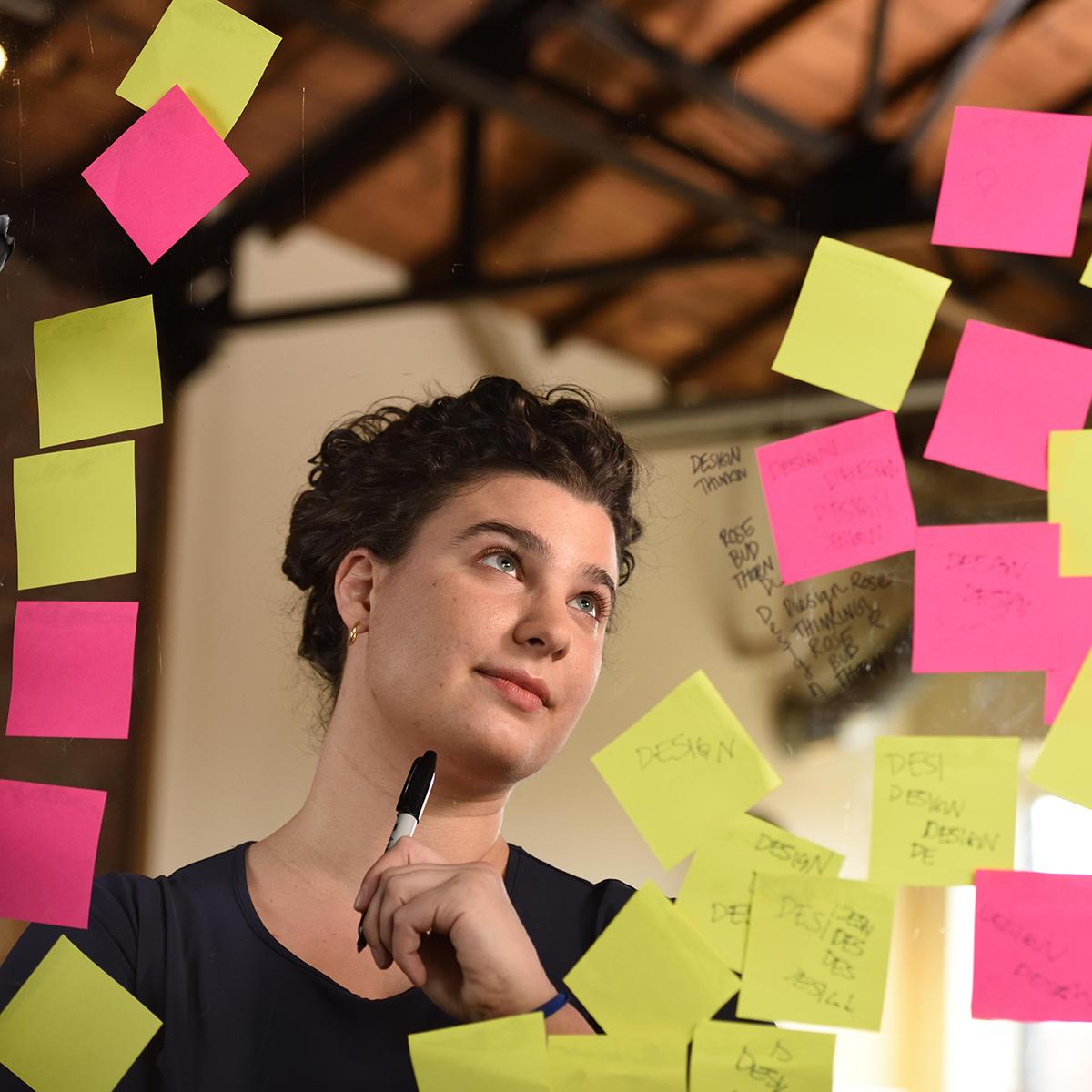 Photo of a young white brunette woman writing on clear glass covered in Post-It notes