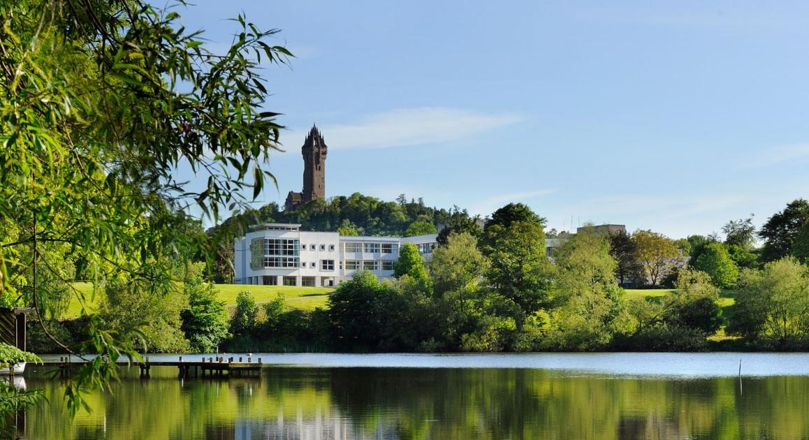 Photo of green forests and a calm lake, 后面有一座白色的建筑和一座高塔:斯特林的斯特林大学, 苏格兰.