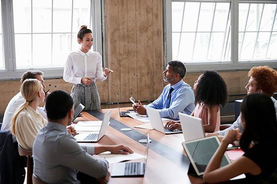 Photo of a woman standing at the head of a conference room table presenting to peers in an office.