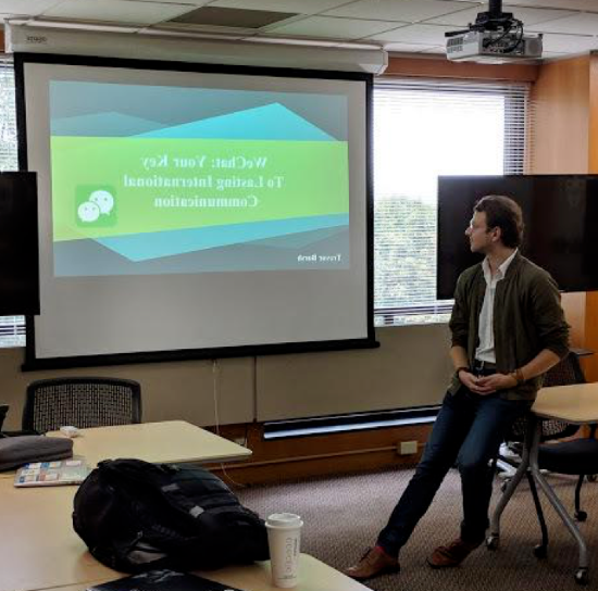 Photo of a PowerPoint title slide (WeChat: Your Key to Lasting International Communication) displayed on a screen in a classroom, with a man leaning against a desk in front of it