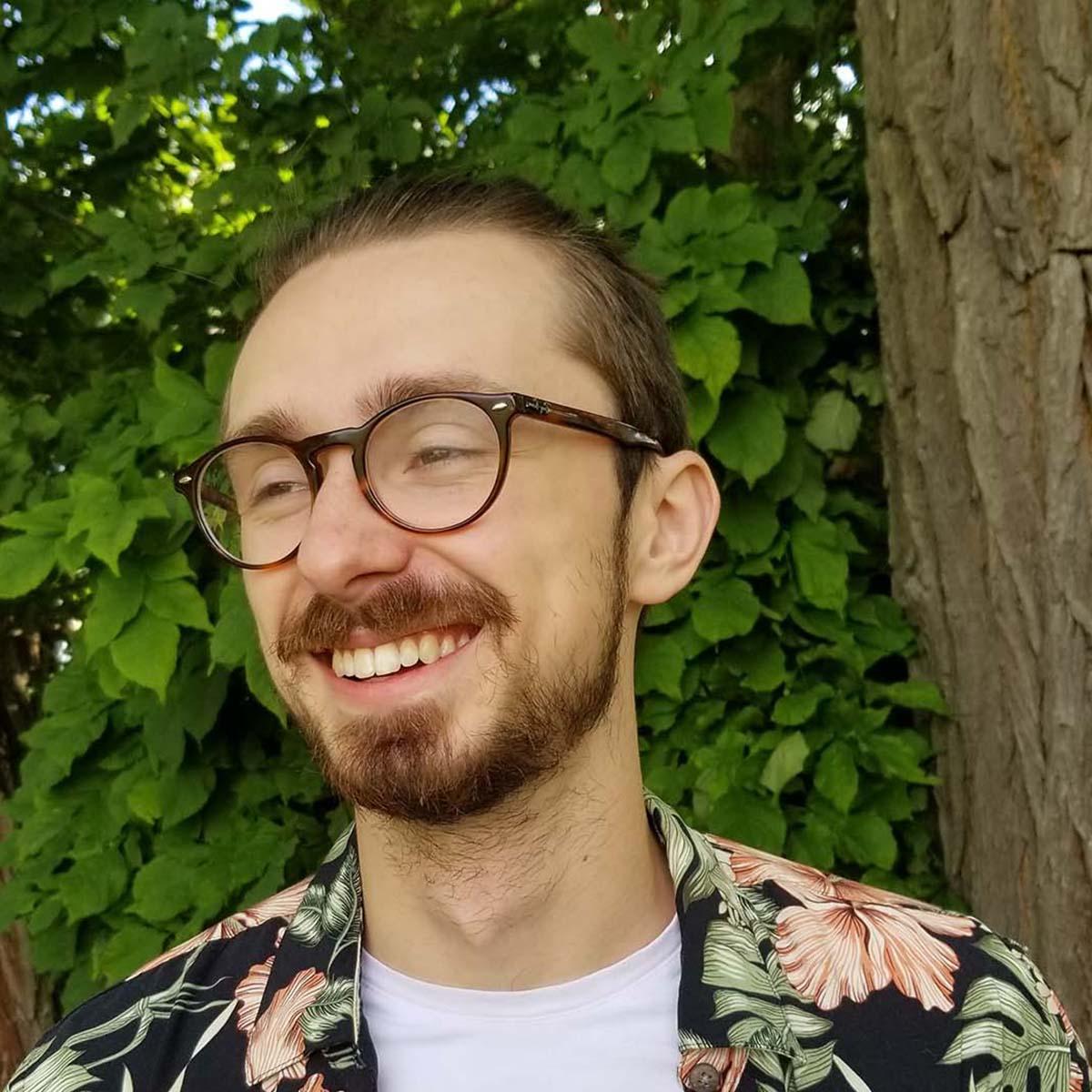 Photo of Carson Custer, 一个戴着眼镜，穿着鲜艳印花衬衫的白人年轻人, smiling in front of greenery