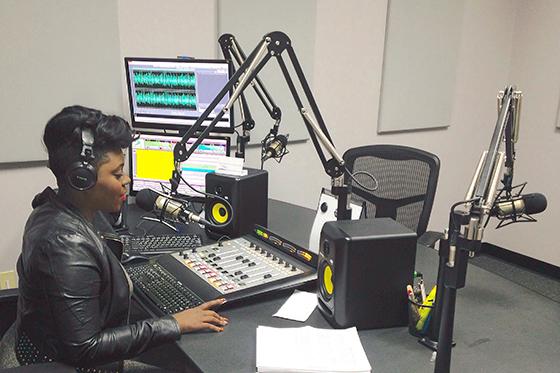 Photo of a woman in a black leather jacket wearing headphones while sitting in a studio with computer monitors and a sound board. 