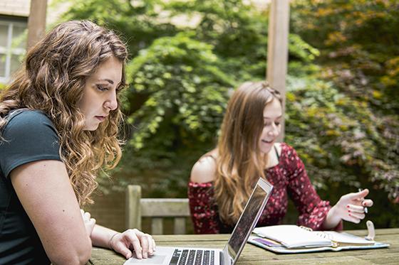 Photo of two Chatham University students sitting outside at a picnic table working on a laptop and writing in a notebook. 