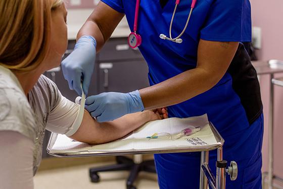 Photo of a nurse in blue scrubs preparing to draw blood from a patient sitting in a chair. 