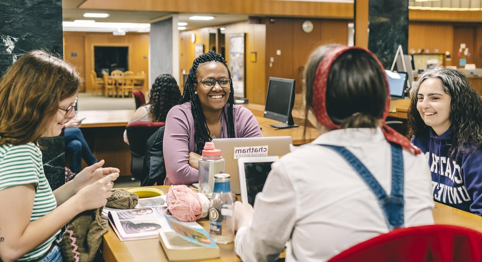 Photo of four female Chatham University students sitting at a table in Jennie Mellon King Library talking with laptops, 笔记本电脑, 和针织. 