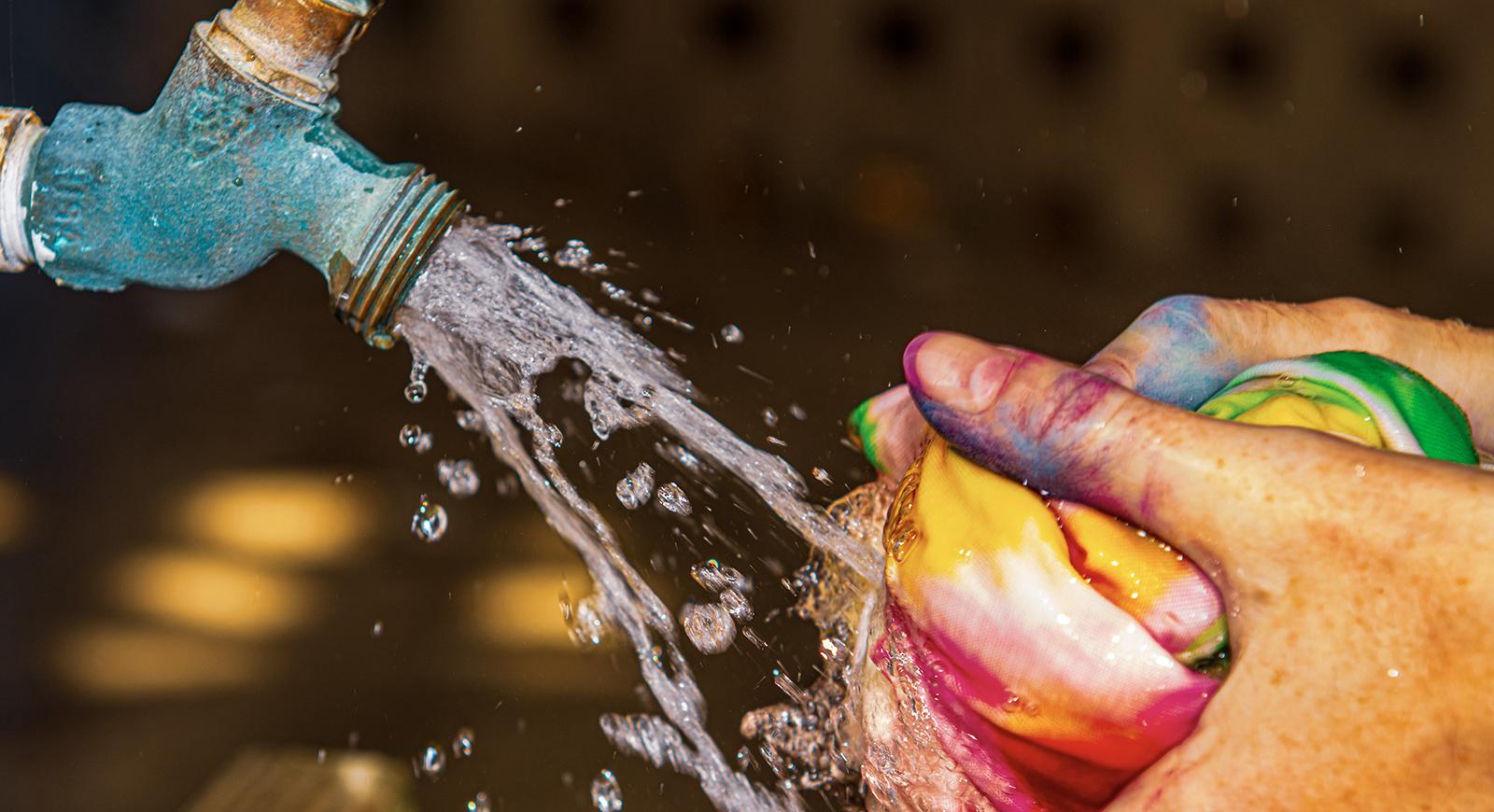 Close-up photo of h和s holding a tie-dyed t-shirt under running water rinsing out excess dye. 