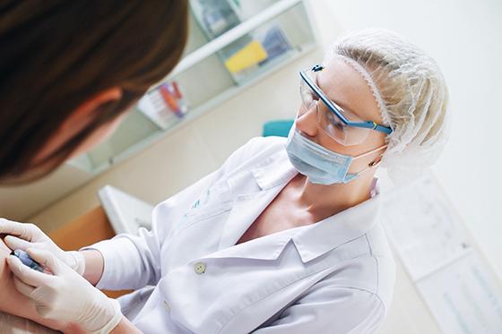 A medical professional wearing a white coat, glasses, and medical mask administers a needle to a patient's arm. 