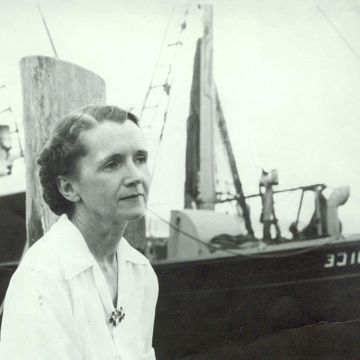 Black 和 white photo of 瑞秋卡森 sitting on a dock, with a pencil 和 paper in her h和 a boat in the background