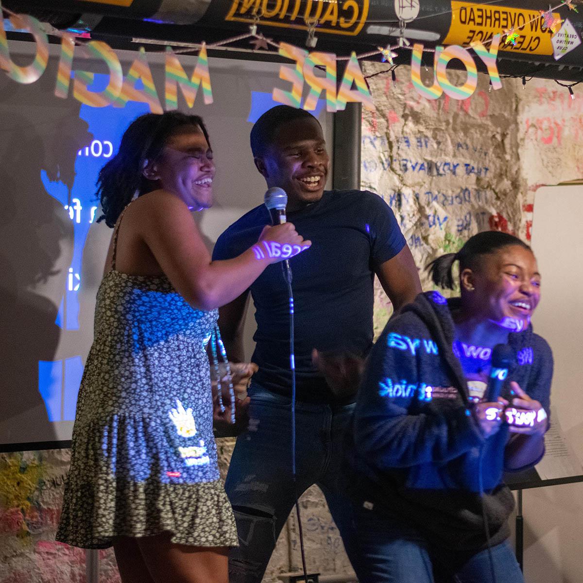 Photo of four Black students laughing and sining on stage with microphones during BIPOC karaoke night. There is a sign behind them that says YOU ARE MAGIC.